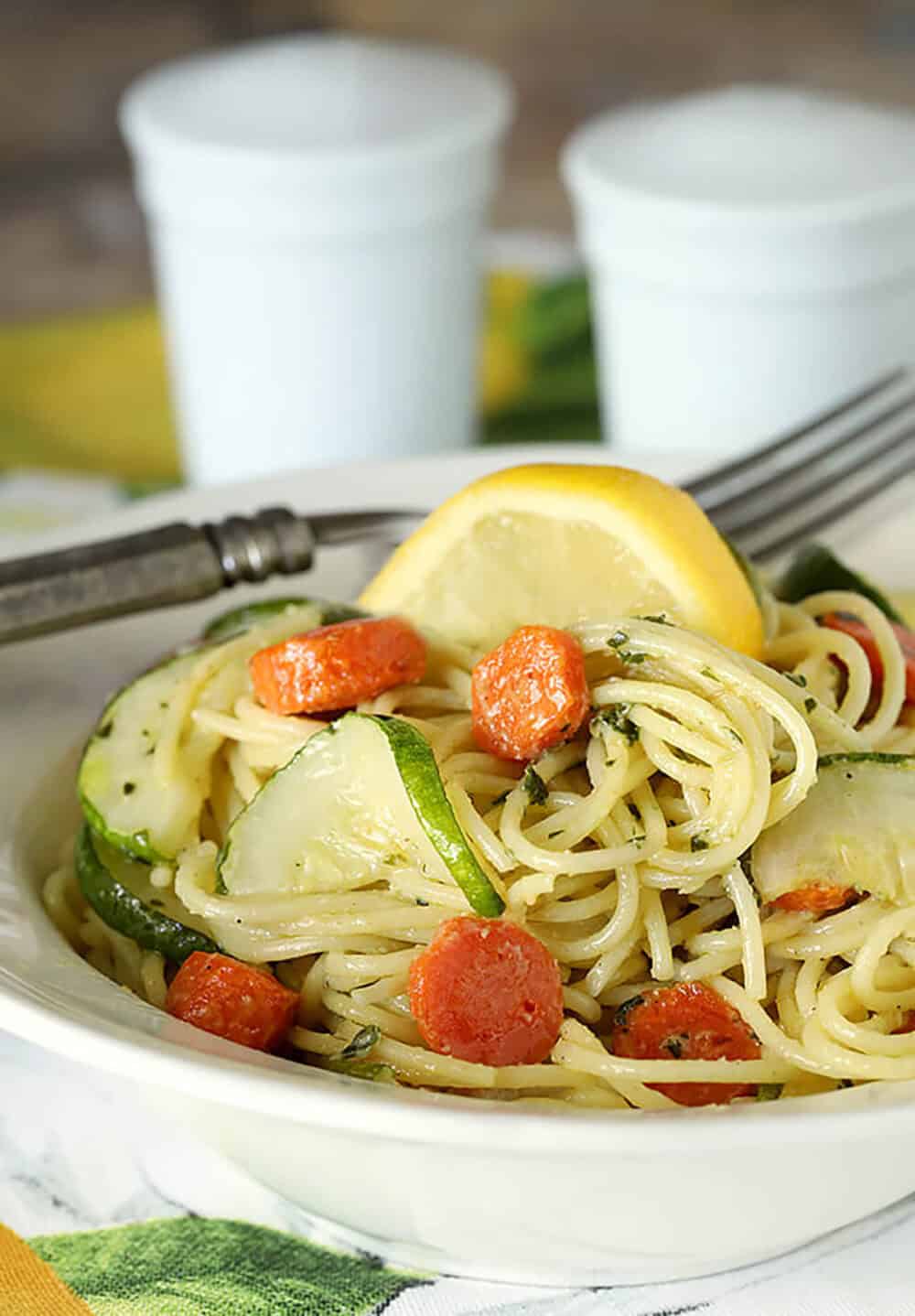 Close-up of garlic pesto pasta with carrots, zucchini, and lemon wedge on top.
