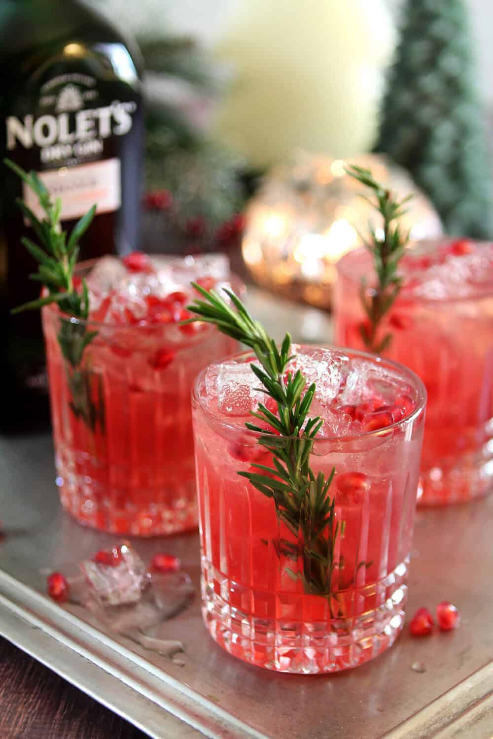 Glasses of Pomegranate and Rosemary Gin Fizz in a lowball cut crystal glass; garnished with a spring of rosemary and pomegranate arils in front of a bottle of Nolets Gin.
