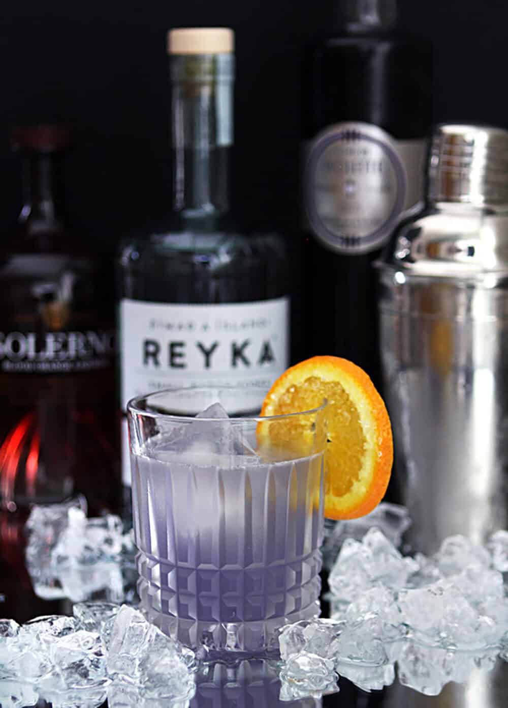 Violet cocktail with orange slice on a glass, surrounded by ice and cocktail ingredients.