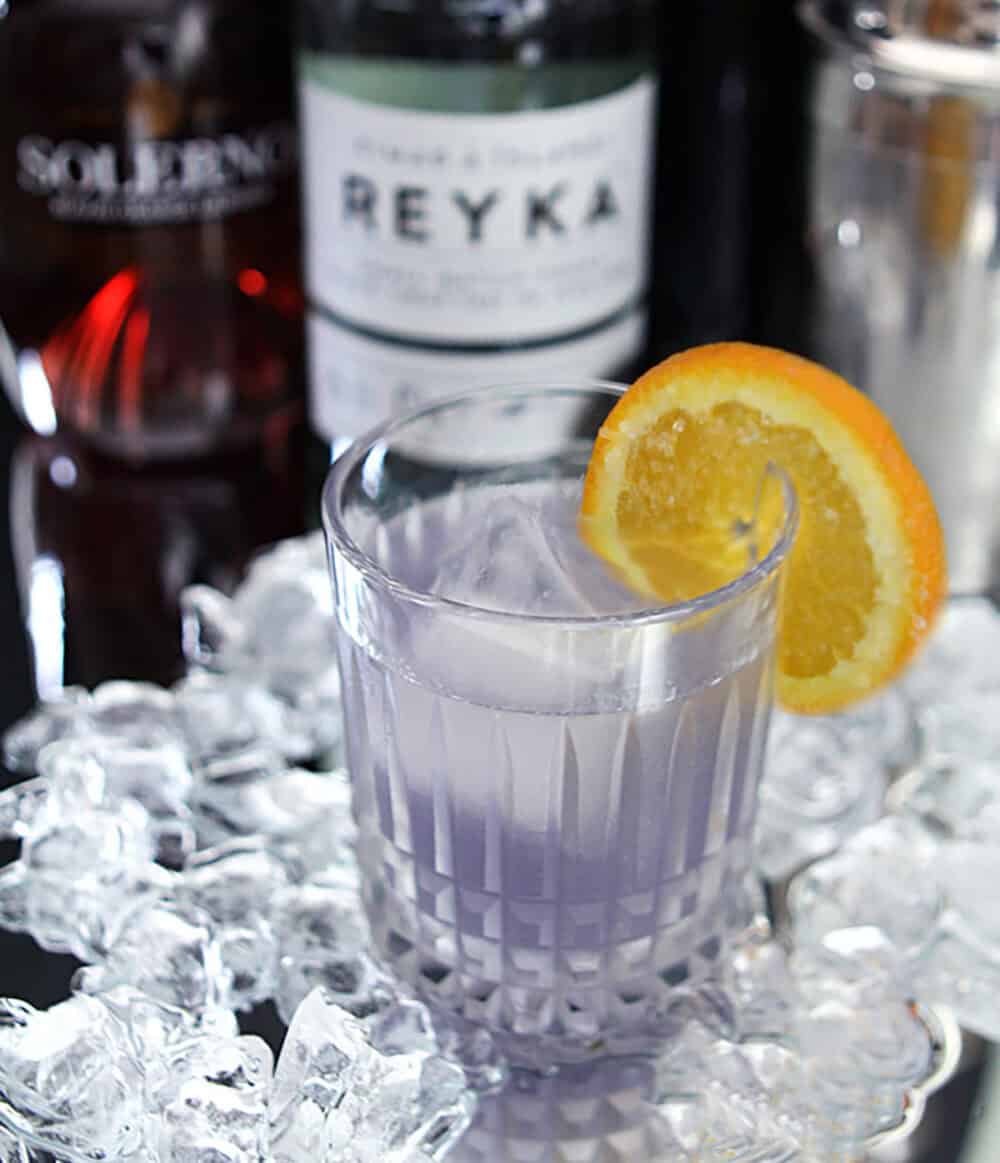 Violet cocktail with orange slice on glass, ice, and liquor bottles in background.
