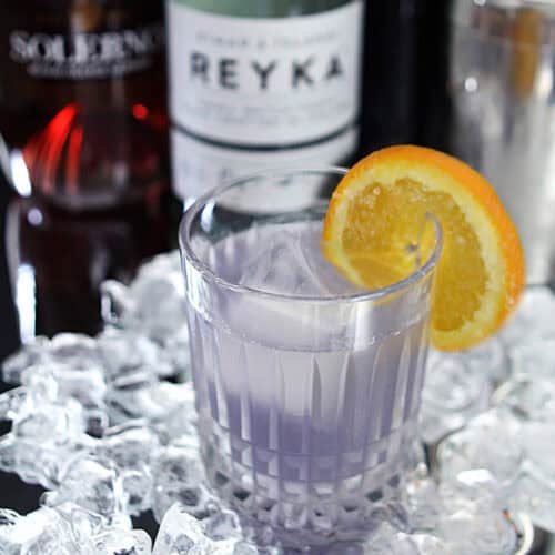 Vodka and Creme de Violette cocktail with orange slice on glass, ice, and liquor bottles in background.